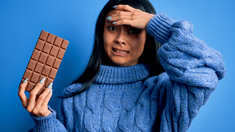 Woman in blue sweater holding a bar of chocolate holding her hand to her forehead in pain