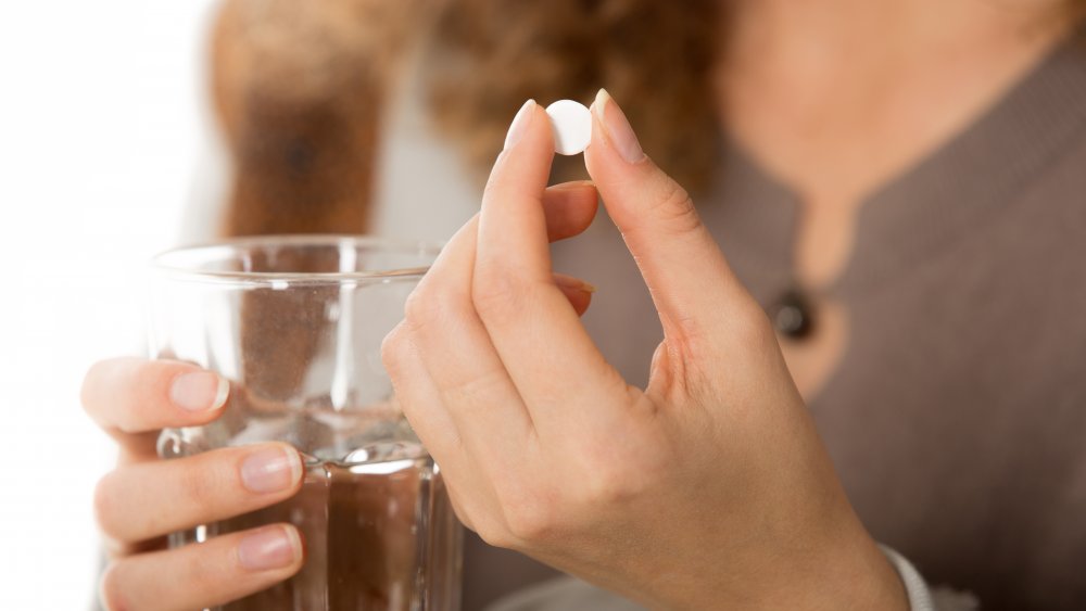 A close-up view photo of female hands holding one white round pill and glass of water