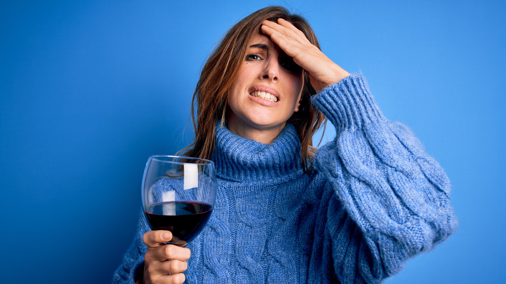 Person in blue sweater grimacing and holding glass of red wine with hand on head