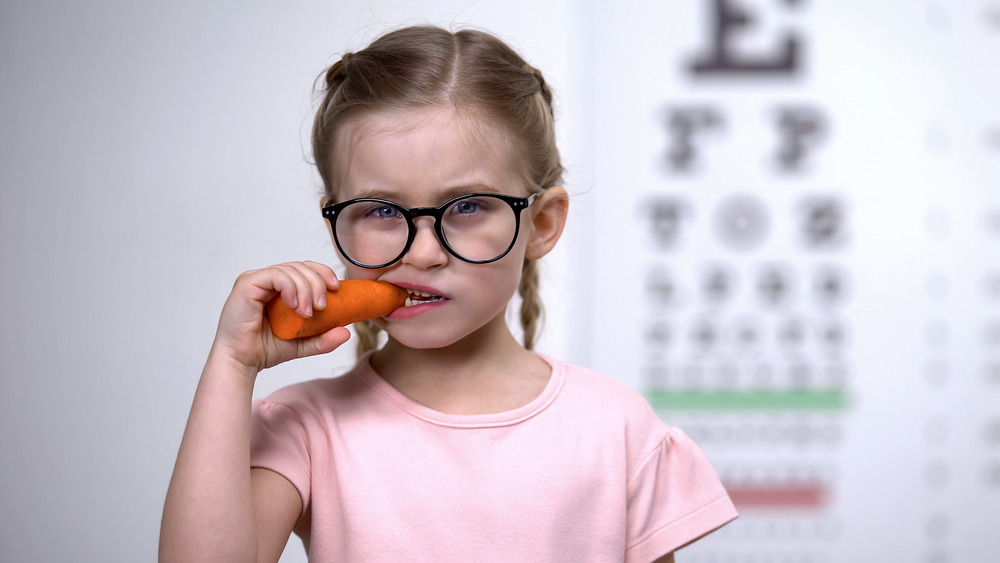 young child wearing glasses, chewing on a carrot, with a vision test in the background 