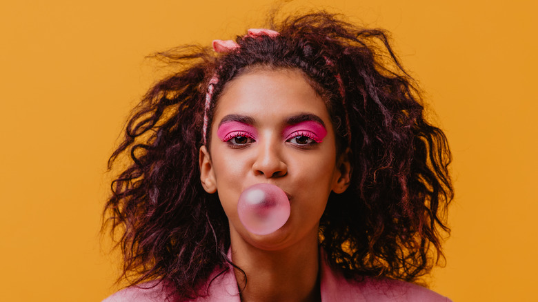 Woman with pink eyeshadow blowing a pink bubble gum bubble on orange background